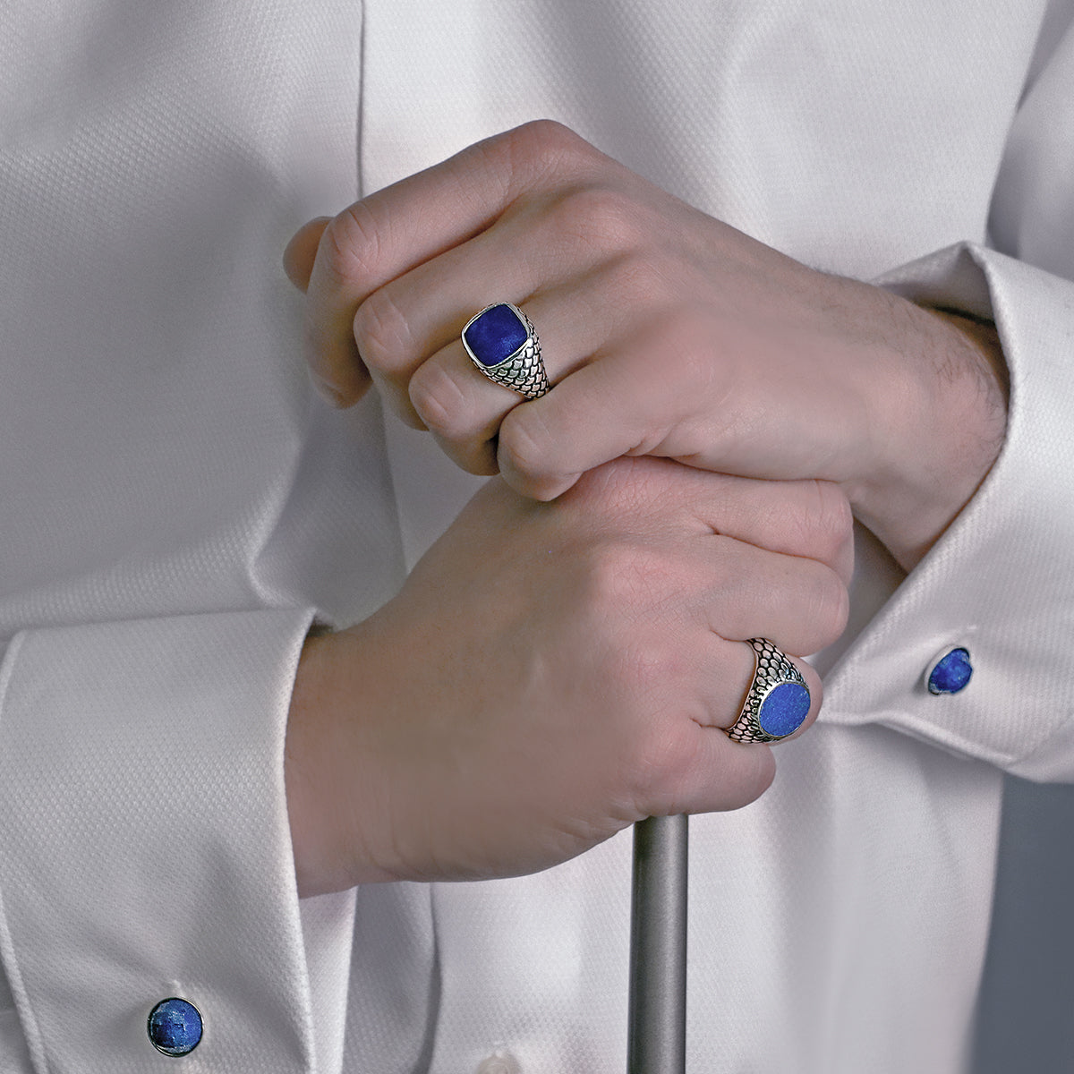 Hands wearing ALBERT M. JEWELS JEWELRY Mod. WSOX00174.TRQ rings with blue gemstones, dressed in a white shirt with matching cufflinks.